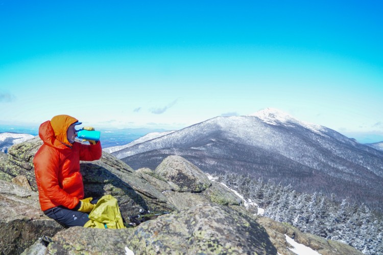 history of puffer jacket: man drinking water in puffy coach with mountains and blue sky in background