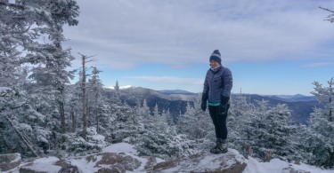 person standing on rock in puffy coat surrounded by snow and open sky