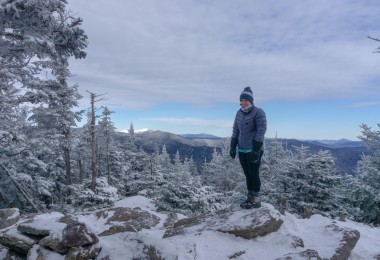 person standing on rock in puffy coat surrounded by snow and open sky