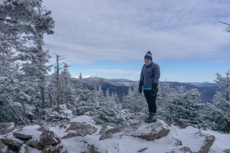 person standing on rock in puffy coat surrounded by snow and open sky