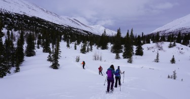 people snowshoeing down a snowy hill with mountains in background