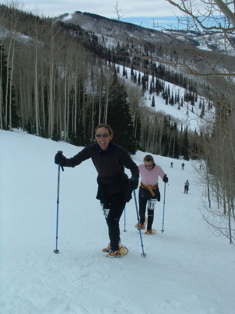 two women going up a hill on snowshoes and poles with racing numbers