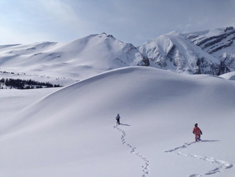 people in distance on snowshoes underneath large hill with snowy mountains in background