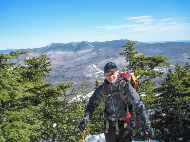 person posing for camera with snowshoes attached to their pack in front of trees and view to mountains beyond