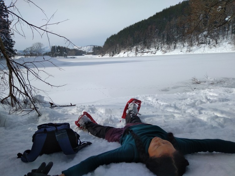 woman laying on back with snowshoes near a lake