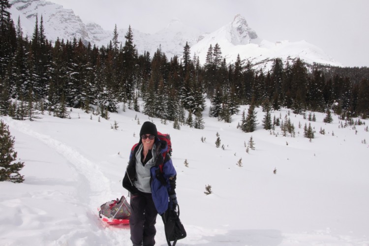 person smiling while carrying gear in front of trees and snowy mountain
