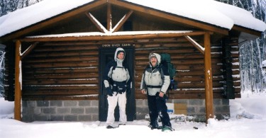 two men standing outside a log cabin in winter