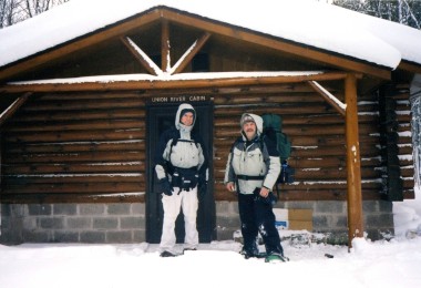 two men standing outside a log cabin in winter