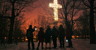 group of people on snowshoes standing beneath illuminated cross at night