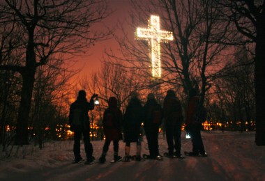 group of people on snowshoes standing beneath illuminated cross at night