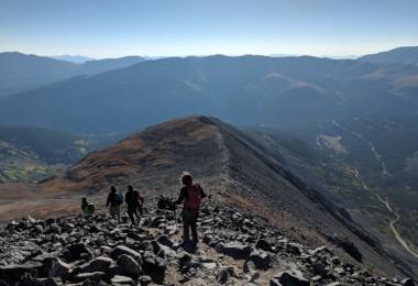people descending mountain summit with trail and blue sky in the background