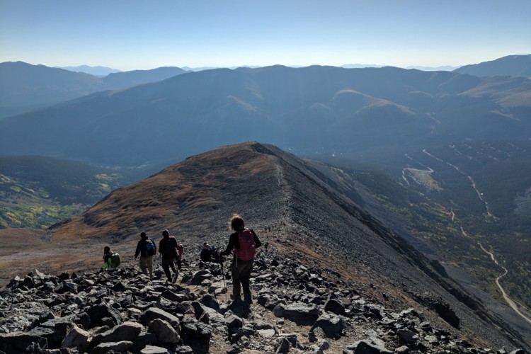people descending mountain summit with trail and blue sky in the background