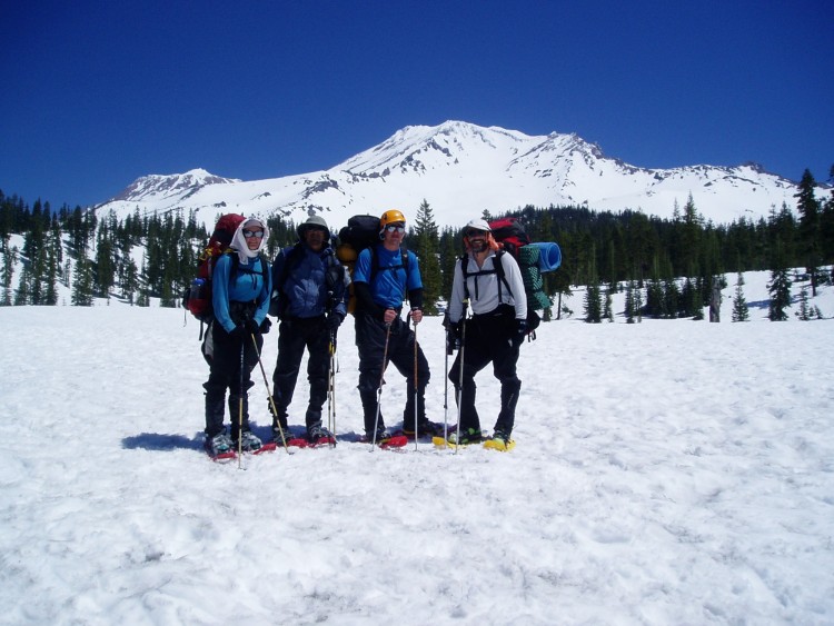 four people with mountaineering equipment on snowshoes with snowy mountains in background