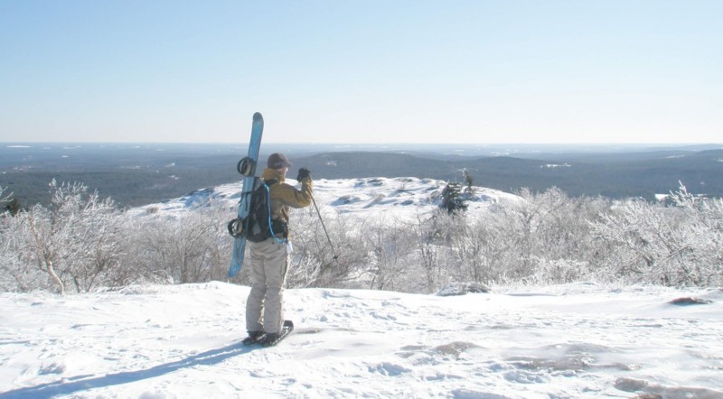 man looking out at view with snowboard in pack and on snowshoes