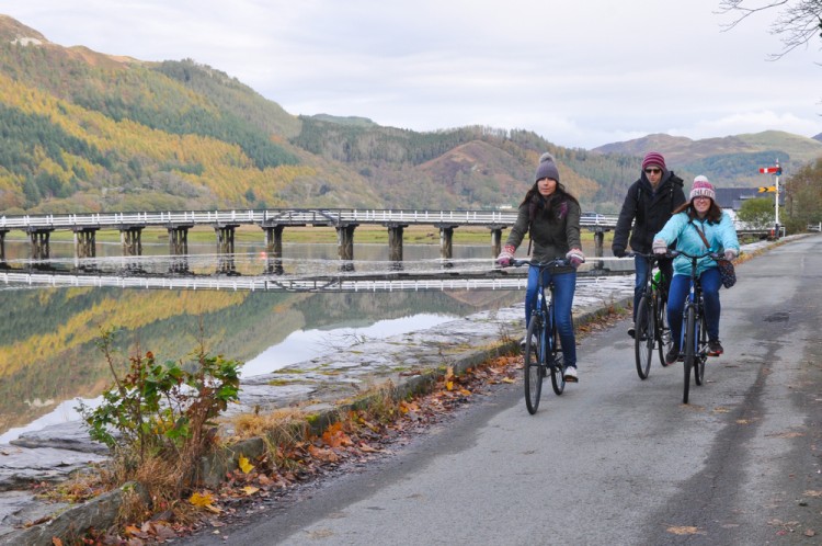 three bikers on trail near river with bridge and fall/winter trees in background