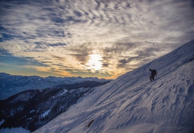 person snowshoeing down a steep mountain with sunset and other peaks in the background