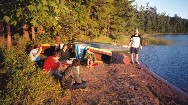 college students sitting down next to canoe near lake