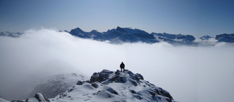 man on rocky ledge surrounded by clouds with blue sky above