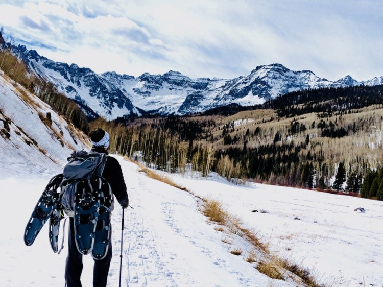 person with multiple pairs of snowshoes on their pack hiking through the mountains